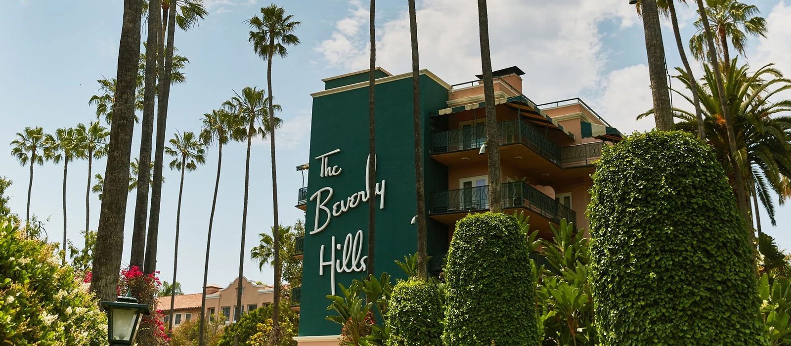 Green sign reading "the beverly hills" on a teal building with balconies, surrounded by palm trees and manicured hedges under a clear sky.