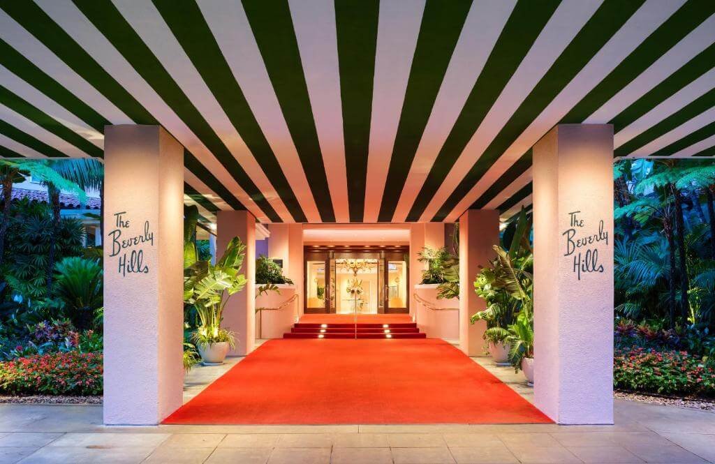 A red carpet entrance to the Beverly Hills hotel, flanked by lush green plants and illuminated by warm lighting under a striped awning, perfect for a celebrity appearance post-microblading session.