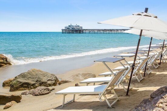 A serene beach setting with a row of white lounge chairs under a large white umbrella, overlooking a calm sea and a distant pier, perfect for relaxing after microblading.